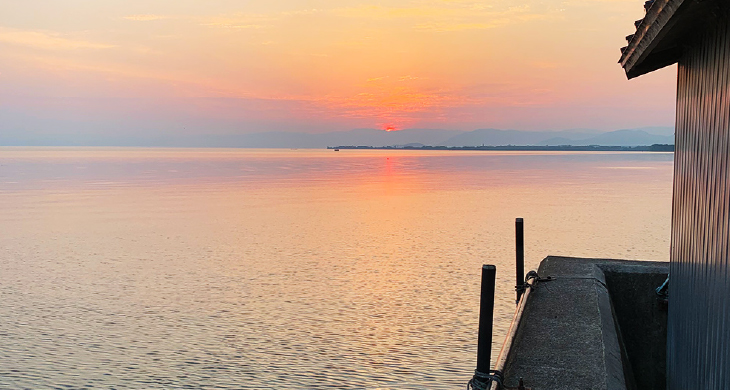 琵琶湖の夕焼けの景色。写真真ん中に稜線に沈みかけている太陽があり、日没で赤く染まっている。