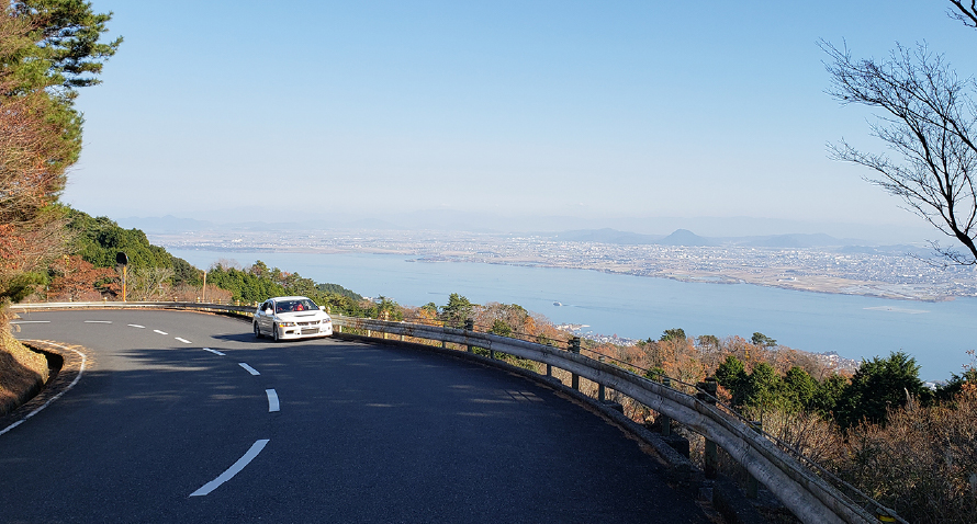 日中に長浜周辺をドライブしている様子。正面からみて左にカーブしている道路を白い自動車が走っている。写真右側のガードレールの奥には水辺を挟んで対岸の景色が見える。