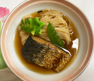 白地に明るい茶色で縁取られた丼に盛られた鯖そうめんの真上からの画像。茶色いだしに黄色いそうめんがあり、その上にさやえんどう1本と焼き鯖の切り身、薬味が乗っている。