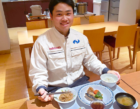 男性職員社員が食堂で定食を食べている様子の画像。明るいブラウンの6人掛けの机と椅子に男性社員が制服を着て座っており、ライトブルーのトレーに乗った定食の横に左手を置き、箸を右手に持ちながら正面を向いて笑顔を向けている。